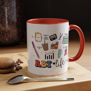 Könnte beinhalten: Eine weiße Keramik-Kaffeetasse mit rotem Rand und Henkel. Die Tasse zeigt eine bunte Illustration verschiedener Symbole, die mit dem Bereich der Verhaltenstherapie zusammenhängen, darunter eine Klemmbrett, ein Diagramm, eine Kaffeetasse und eine Stoppuhr. Der Text "RBT life" ist in bunten Buchstaben am Boden der Tasse geschrieben.