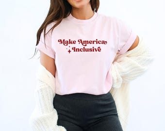 Può includere: Una maglietta rosa con il testo "Make America Inclusive" in lettere rosse con un design a stella.