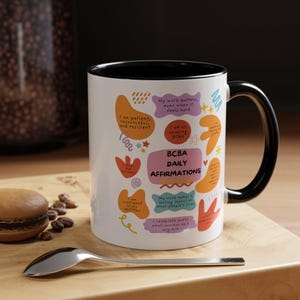 May include: White ceramic mug with a black rim and handle. The mug features colorful abstract shapes with text that reads "BCBA Daily Affirmations". The text includes phrases like "I am patient, resourceful, and resilient" and "My work matters, even when it feels hard".