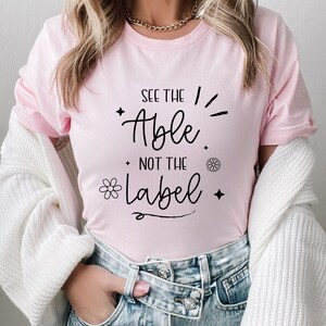 May include: A light pink t-shirt with the text "SEE THE ABLE NOT THE LABEL" in black script. The design includes a flower, stars, and radiating lines. The shirt is part of a casual wear collection.