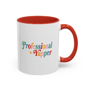 Könnte beinhalten: Weiße Keramiktasse mit rotem Rand und Henkel. Die Tasse hat eine bunte Grafik mit der Aufschrift "Professional Yapper" mit einem Glitzern.