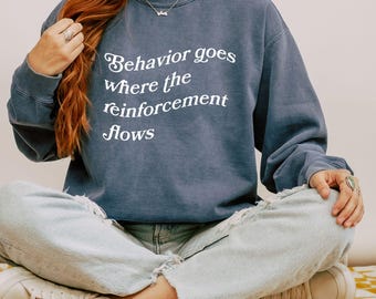 Puede incluir: Una sudadera azul marino con el texto "Behavior goes where the reinforcement flows" impreso en blanco en la parte delantera.