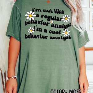 K&ouml;nnte beinhalten: Ein gr&uuml;nes T-Shirt mit dem Text "I'm not like a regular behavior analyst I'm a cool behavior analyst" in Schwarz mit wei&szlig;en G&auml;nsebl&uuml;mchen um den Text herum. Der Text "COLOR - MOSS" befindet sich am unteren Rand des Shirts.