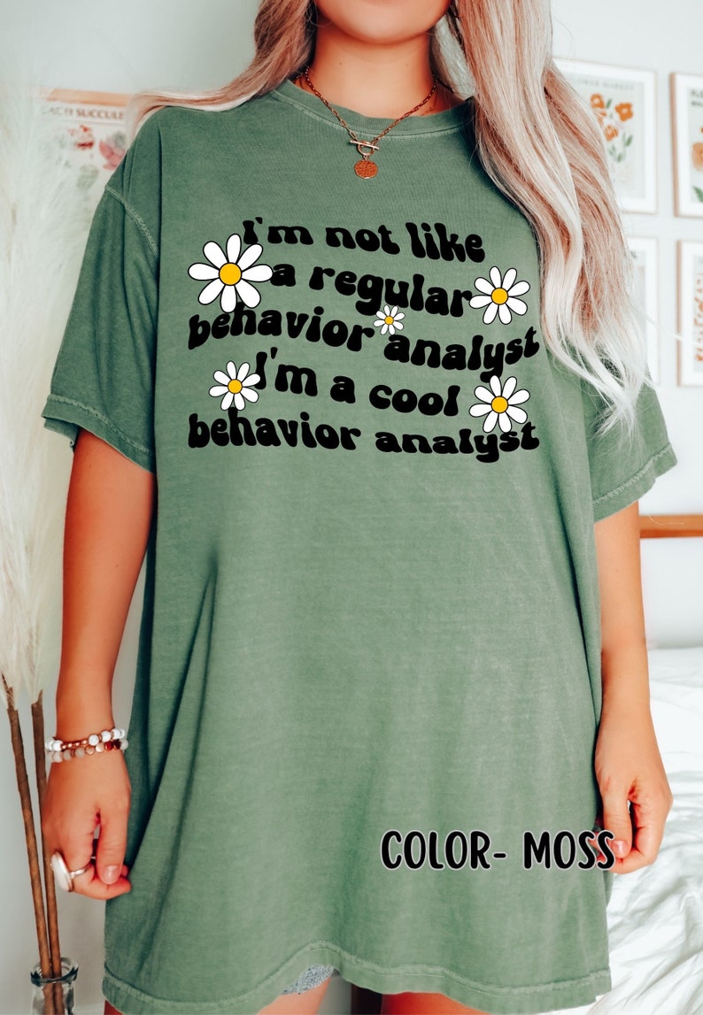K&ouml;nnte beinhalten: Ein gr&uuml;nes T-Shirt mit dem Text "I'm not like a regular behavior analyst I'm a cool behavior analyst" in Schwarz mit wei&szlig;en G&auml;nsebl&uuml;mchen um den Text herum. Der Text "COLOR - MOSS" befindet sich am unteren Rand des Shirts.