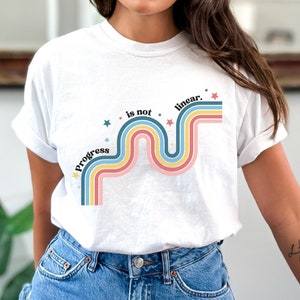 Pu&ograve; includere: Maglietta bianca con un design arcobaleno colorato e il testo "Progress is not linear."