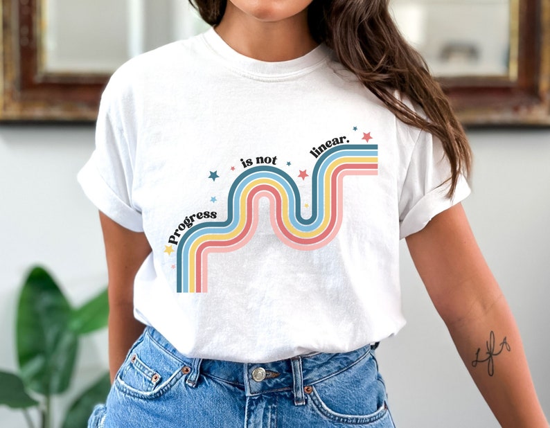 Pu&ograve; includere: Maglietta bianca con un design arcobaleno colorato e il testo "Progress is not linear."
