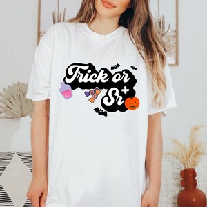 Puede incluir: Camiseta blanca con un gr&aacute;fico negro que dice "Trick or Sr+" con una botella de poci&oacute;n morada, caramelos y una calabaza.