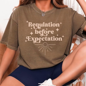 May include: A brown short-sleeve crewneck t-shirt featuring the text "Regulation before Expectation" printed in a light beige colour. The design includes small four-pointed star icons and a sunburst graphic positioned below the text.