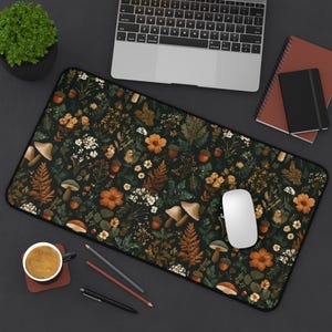 May include: A rectangular desk mat featuring a dark green background with a botanical pattern of mushrooms, flowers, ferns, and acorns in shades of orange, brown, and white. A white computer mouse rests on the mat.