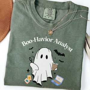 Op de afbeelding: Een mosgroen t-shirt met een witte afbeelding van een geest die een bellenblaas en een ijskoffie vasthoudt, met een laptop en kleine iconen erbij. De tekst luidt Boo-Havior Analyst. Het shirt heeft een Comfort Colors label in de nek.
