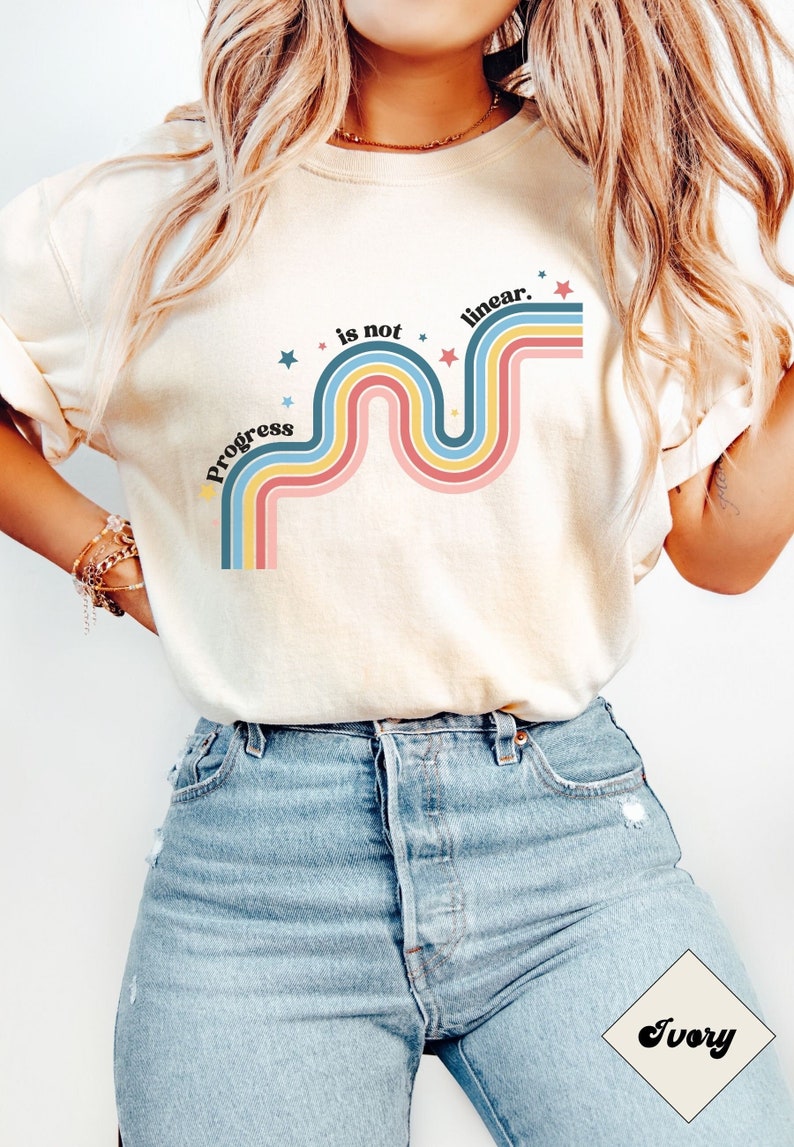 Pu&ograve; includere: Una maglietta bianca con un design grafico di una linea ondulata arcobaleno con il testo "Progress is not linear." in nero. La grafica &egrave; circondata da piccole stelle bianche.
