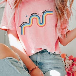 Pu&ograve; includere: Una maglietta rosa con un design arcobaleno retr&ograve; e il testo "Progress is not linear." L'arcobaleno &egrave; composto da strisce rosse, arancioni, gialle, blu e verdi. Ci sono piccole stelle bianche sparse intorno al design.