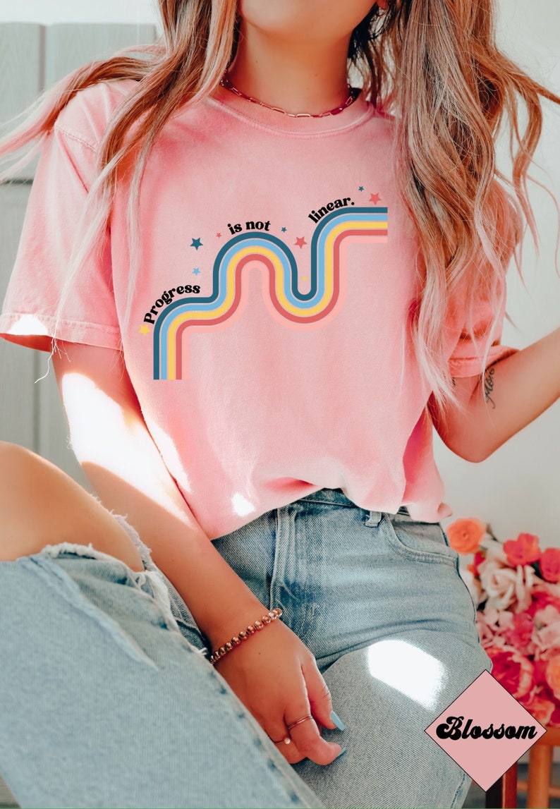 Pu&ograve; includere: Una maglietta rosa con un design arcobaleno retr&ograve; e il testo "Progress is not linear." L'arcobaleno &egrave; composto da strisce rosse, arancioni, gialle, blu e verdi. Ci sono piccole stelle bianche sparse intorno al design.