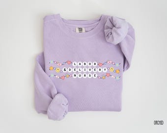 Könnte beinhalten: Ein lavendelfarbener Pullover mit dem Text "LABOR & DELIVERY NURSE" in weißen Buchstaben, umgeben von bunten Herzen, Sternen und Smiley-Gesichtern.