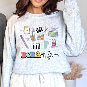 Puede incluir: Una sudadera gris claro con un diseño gráfico colorido. El diseño incluye varios iconos como una calculadora, una taza de café y un bolígrafo, junto con el texto "BCBA life" en una fuente lúdica. La sudadera es de estilo casual.