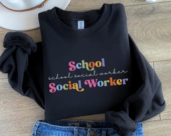 Puede incluir: Sudadera negra con las palabras "School school social worker Social Worker" en letras coloridas de estilo retro. La sudadera se combina con vaqueros azules y un sombrero blanco.