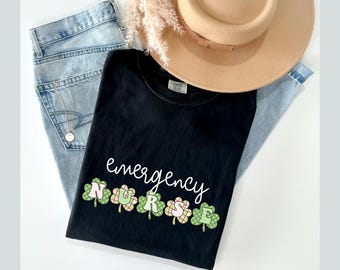 May include: Black t-shirt with the text "emergency nurse" in white script. The word "nurse" is spelled out with green shamrock-shaped letters.