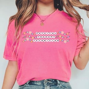 May include: Pink t-shirt with a graphic of a string of lights with the words "PHYSICAL THERAPY TECHNICIAN" on it.