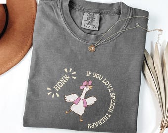 May include: A gray Comfort Colors t-shirt featuring a cartoon goose wearing a pink cowboy hat and scarf. The text reads "HONK IF YOU LOVE SPEECH THERAPY" in a circular design. A gold necklace with a pendant is also visible.