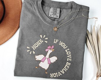 May include: Dark gray Comfort Colors t-shirt with a cartoon goose wearing a pink cowboy hat and scarf. The shirt has the text "HONK IF YOU LOVE BEHAVIOR" in a circular design. A gold necklace is on the shirt.