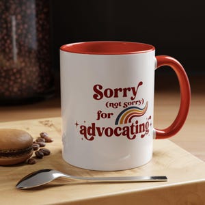May include: White ceramic mug with a red interior and handle. The mug features the text "Sorry (not sorry) for advocating" in a retro font with a rainbow graphic. The mug is on a wooden surface with a spoon and a pastry.
