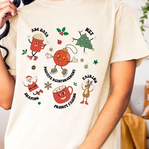 May include: A cream coloured t-shirt with a Christmas themed design featuring cartoon characters and text that says "ABC Data", "Net", "Positive Reinforcement", "Shaping", "Pairing", "Prompt Fading".