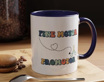 May include: White ceramic mug with a navy blue interior and handle. The mug features the text "FINE MOTOR PROMOTOR" in colourful, retro-style lettering, along with a dotted line, heart, and scissors graphic. A spoon and a macaron are on a wooden surface.