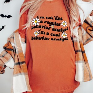K&ouml;nnte beinhalten: Ein orangefarbenes T-Shirt mit dem Text "I'm not like a regular behavior analyst I'm a cool behavior analyst" in schwarzer Schrift mit wei&szlig;en G&auml;nsebl&uuml;mchen um den Text herum. Der Text ist in einem Retro-Schriftart-Stil. Das T-Shirt tr&auml;gt ein Model, das ein kariertes Hemd tr&auml;gt.