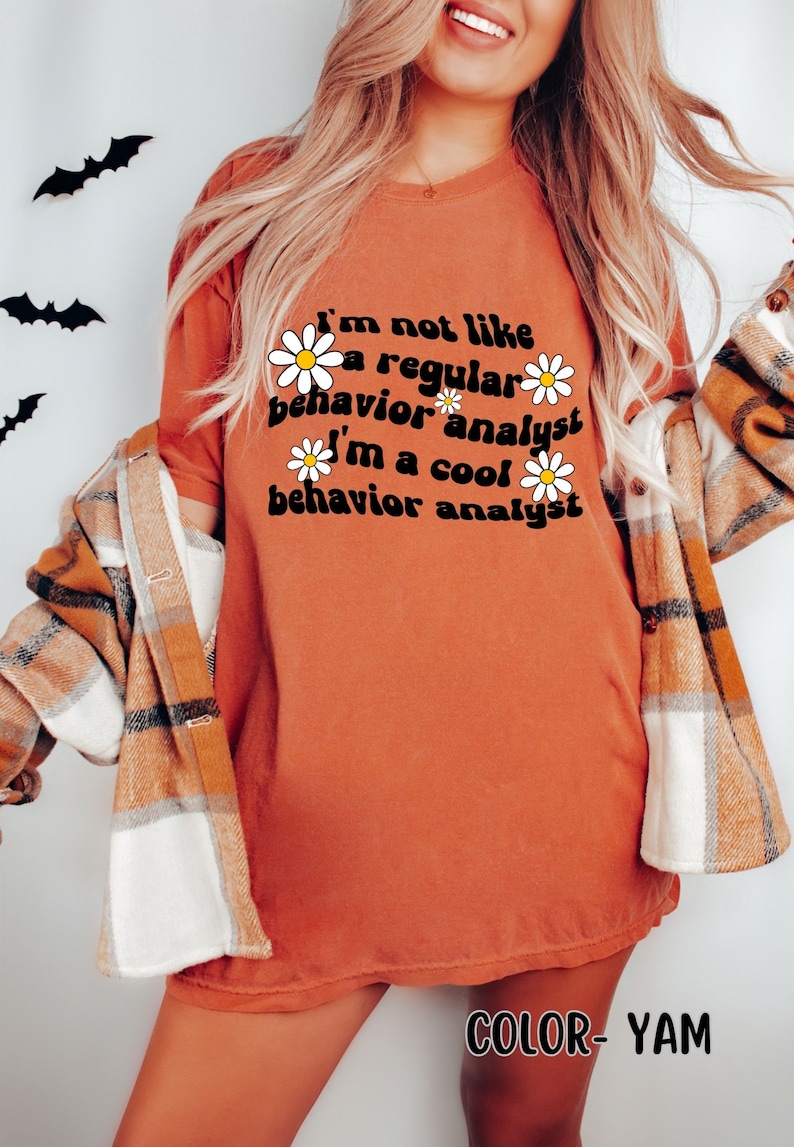 K&ouml;nnte beinhalten: Ein orangefarbenes T-Shirt mit dem Text "I'm not like a regular behavior analyst I'm a cool behavior analyst" in schwarzer Schrift mit wei&szlig;en G&auml;nsebl&uuml;mchen um den Text herum. Der Text ist in einem Retro-Schriftart-Stil. Das T-Shirt tr&auml;gt ein Model, das ein kariertes Hemd tr&auml;gt.