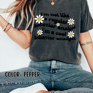 K&ouml;nnte beinhalten: Ein dunkelgraues T-Shirt mit einer Retro-Grafik, auf der "Ich bin nicht wie ein normaler Verhaltensanalytiker Ich bin ein cooler Verhaltensanalytiker" steht, mit wei&szlig;en G&auml;nsebl&uuml;mchen um den Text herum. Der Text ist schwarz und die G&auml;nsebl&uuml;mchen sind wei&szlig; mit gelben Zentren. Das Shirt wird von einer Person mit langen blonden Haaren getragen. Der Text "COLOR- PEPPER" ist am unteren Rand des Bildes sichtbar.