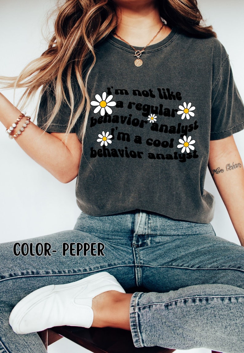 K&ouml;nnte beinhalten: Ein dunkelgraues T-Shirt mit einer Retro-Grafik, auf der "Ich bin nicht wie ein normaler Verhaltensanalytiker Ich bin ein cooler Verhaltensanalytiker" steht, mit wei&szlig;en G&auml;nsebl&uuml;mchen um den Text herum. Der Text ist schwarz und die G&auml;nsebl&uuml;mchen sind wei&szlig; mit gelben Zentren. Das Shirt wird von einer Person mit langen blonden Haaren getragen. Der Text "COLOR- PEPPER" ist am unteren Rand des Bildes sichtbar.