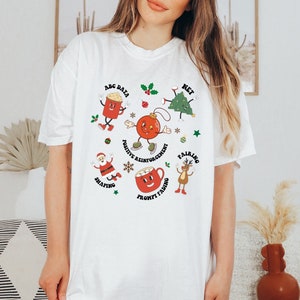 May include: White t-shirt with a Christmas theme featuring cartoon characters and text related to positive reinforcement, shaping, pairing, prompt fading, ABC data, and net.