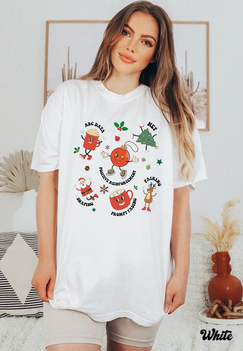 May include: White t-shirt with a Christmas theme featuring cartoon characters and text related to positive reinforcement, shaping, pairing, prompt fading, ABC data, and net.