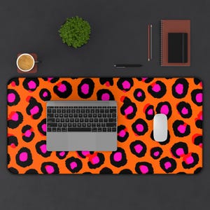 May include: An orange desk mat with a leopard print design in black and pink. A silver laptop keyboard and white mouse are on the mat. A cup of coffee, a small plant, a pen, and a notebook are also on the dark surface.