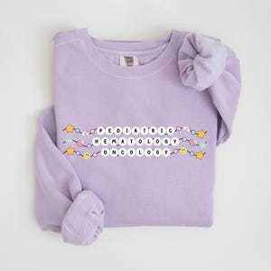 Może przedstawiać: Lavenדרowa bluza z napisem "PEDIATRIC HEMATOLOGY ONCOLOGY" napisanym w postaci kolorowej koralikowej girlandy.