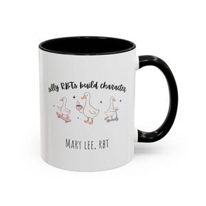 Könnte beinhalten: Weißer Keramikbecher mit schwarzem Innenraum und Griff. Der Becher zeigt den Text "silly RBTs build character" und den Namen "MARY LEE, RBT". Das Design umfasst Cartoon-Enten auf einem Skateboard, die eine Tasse halten und Roller fahren.