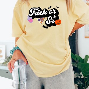 Puede incluir: Una camiseta amarilla con un gr&aacute;fico negro que dice "Trick or Sr+" con ilustraciones de tem&aacute;tica de Halloween.
