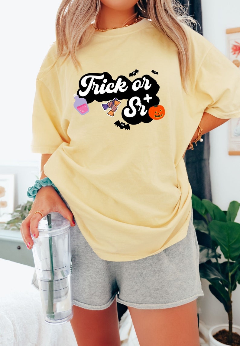 Puede incluir: Una camiseta amarilla con un gr&aacute;fico negro que dice "Trick or Sr+" con ilustraciones de tem&aacute;tica de Halloween.