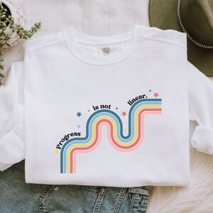 May include: White sweatshirt with a rainbow design and the text "Progress is not linear." in black. The rainbow is made up of pink, yellow, blue, and teal stripes.