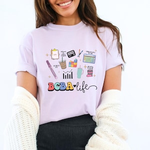 Puede incluir: Una camiseta de color lila claro con un diseño gráfico colorido que presenta varios iconos y el texto "BCBA life". El diseño incluye un bolígrafo, una taza de café, una calculadora y otras imágenes relacionadas. La camiseta está hecha de un material suave.