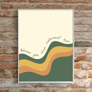 May include: A rectangular wall art print in a thin silver frame featuring a cream background and wavy horizontal bands in dark green, mustard yellow, and terracotta orange. The black cursive text reads, behavior goes where reinforcement flows.
