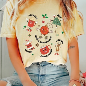 May include: A yellow t-shirt with a Christmas-themed design featuring various cartoon characters and text related to positive reinforcement techniques. The text includes "ABC Data", "Net", "Positive Reinforcement", "Pairing", "Shaping", and "Prompt Fading".