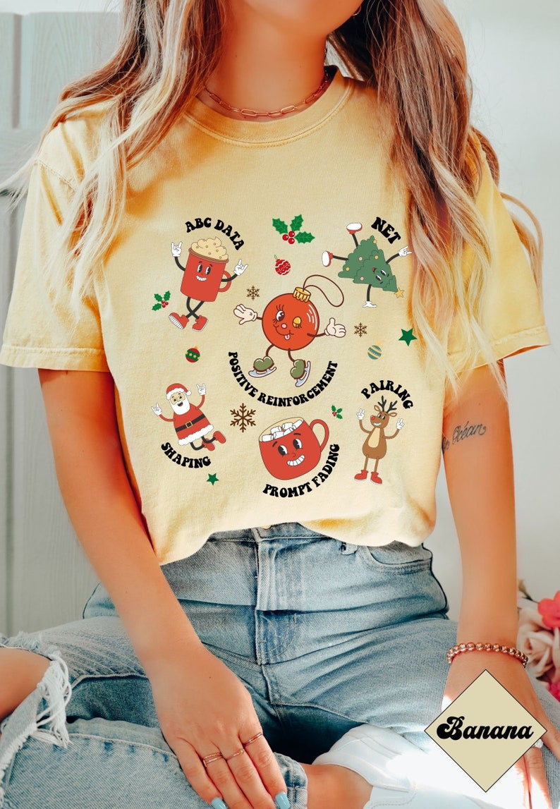 May include: A yellow t-shirt with a Christmas-themed design featuring various cartoon characters and text related to positive reinforcement techniques. The text includes "ABC Data", "Net", "Positive Reinforcement", "Pairing", "Shaping", and "Prompt Fading".