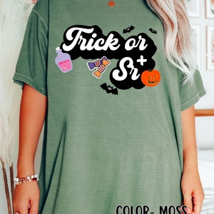 Puede incluir: Una camiseta verde musgo con el texto "Trick or Sr+" en una fuente retro. La camiseta presenta una nube negra con una botella de poci&oacute;n morada, caramelos, murci&eacute;lagos y una calabaza de Halloween.