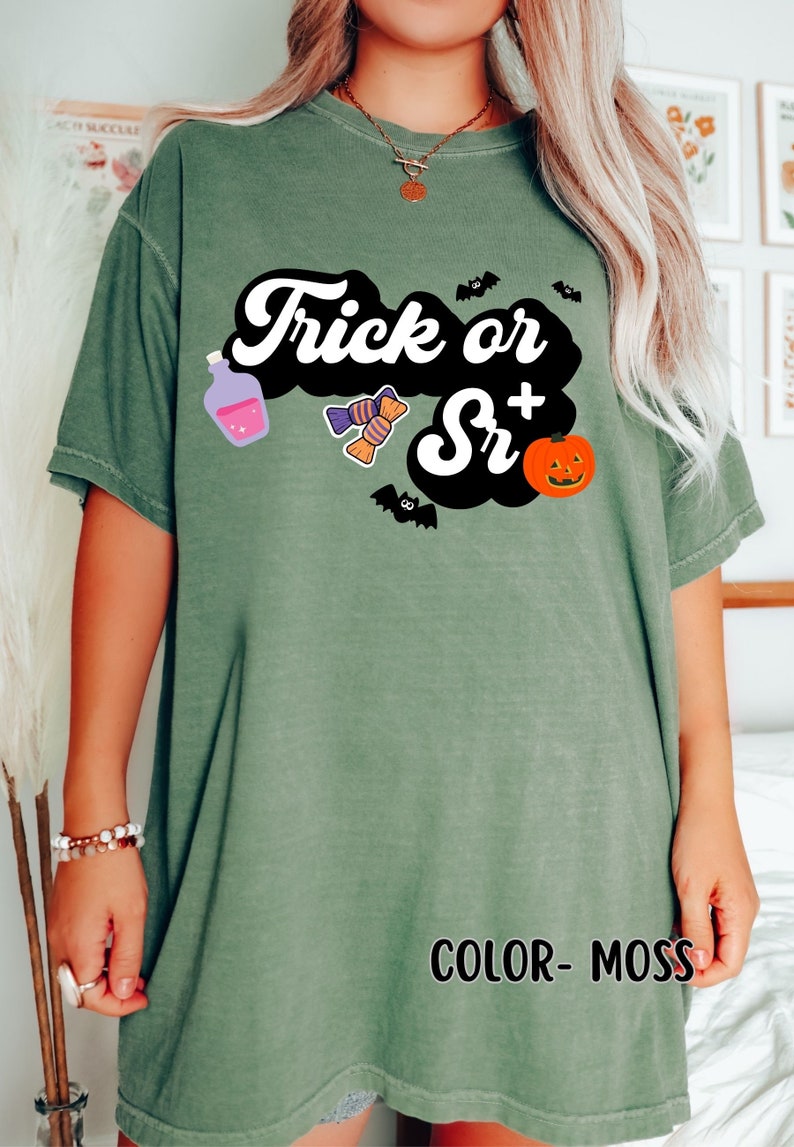 Puede incluir: Una camiseta verde musgo con el texto "Trick or Sr+" en una fuente retro. La camiseta presenta una nube negra con una botella de poci&oacute;n morada, caramelos, murci&eacute;lagos y una calabaza de Halloween.