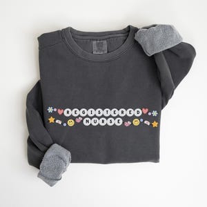 Könnte beinhalten: Ein dunkelgrauer Pullover mit den Worten "REGISTERED NURSE" in weißen Buchstaben auf weißen Kreisen mit bunten Verzierungen.