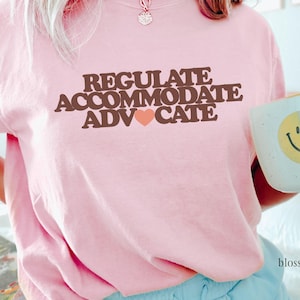 Puede incluir: Sudadera rosa claro con las palabras "REGULATE ACCOMMODATE ADVOCATE" en letras marrones y un pequeño corazón. Una taza blanca con una carita sonriente amarilla se sostiene en la mano.