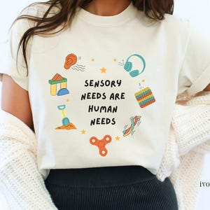 May include: Ivory-colored t-shirt with the text "Sensory Needs Are Human Needs" surrounded by colorful illustrations of sensory items, including headphones, a fidget spinner, and a building block toy.