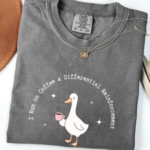Puede incluir: Camiseta gris con cuello redondo. La camiseta presenta un gráfico de un pato blanco sosteniendo una taza de café rosa, con el texto "I run on Coffee & Differential Reinforcement". También es visible un collar plateado con un colgante.