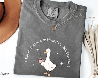 May include: Pepper-colored t-shirt with a round neck. The shirt features a graphic of a white duck holding a pink coffee cup, with the text "I run on Coffee & Differential Reinforcement". A silver necklace with a pendant is also visible.
